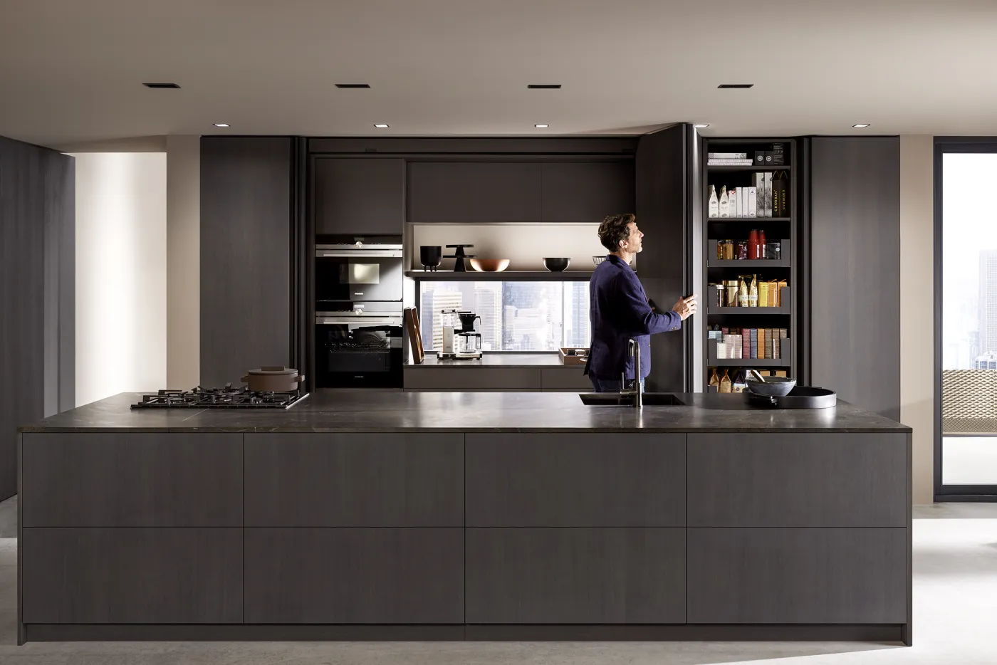 Dunkle Küche mit Kochinsel, dahinter eine Schrankwand mit geöffneten Pocket Doors: Backöfen, beleuchtete Arbeitsnische mit Kaffeemaschine und Vorratsschrank